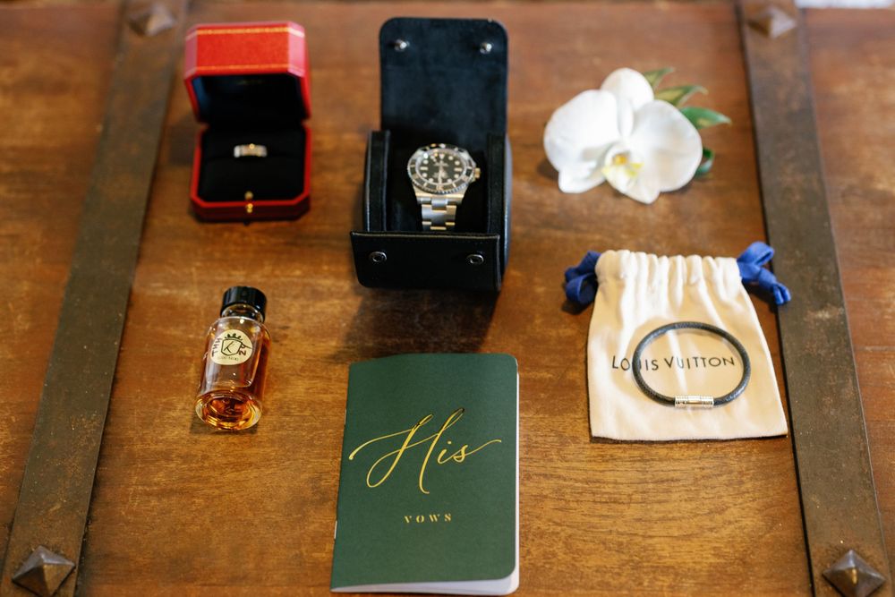 Wedding flat lay with watch, rings, Louis Vuitton pouch, cologne, and His vow book.