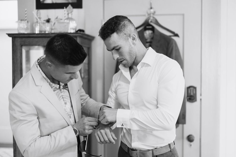 Groomsman helping groom button his suit jacket while getting ready for wedding ceremony.