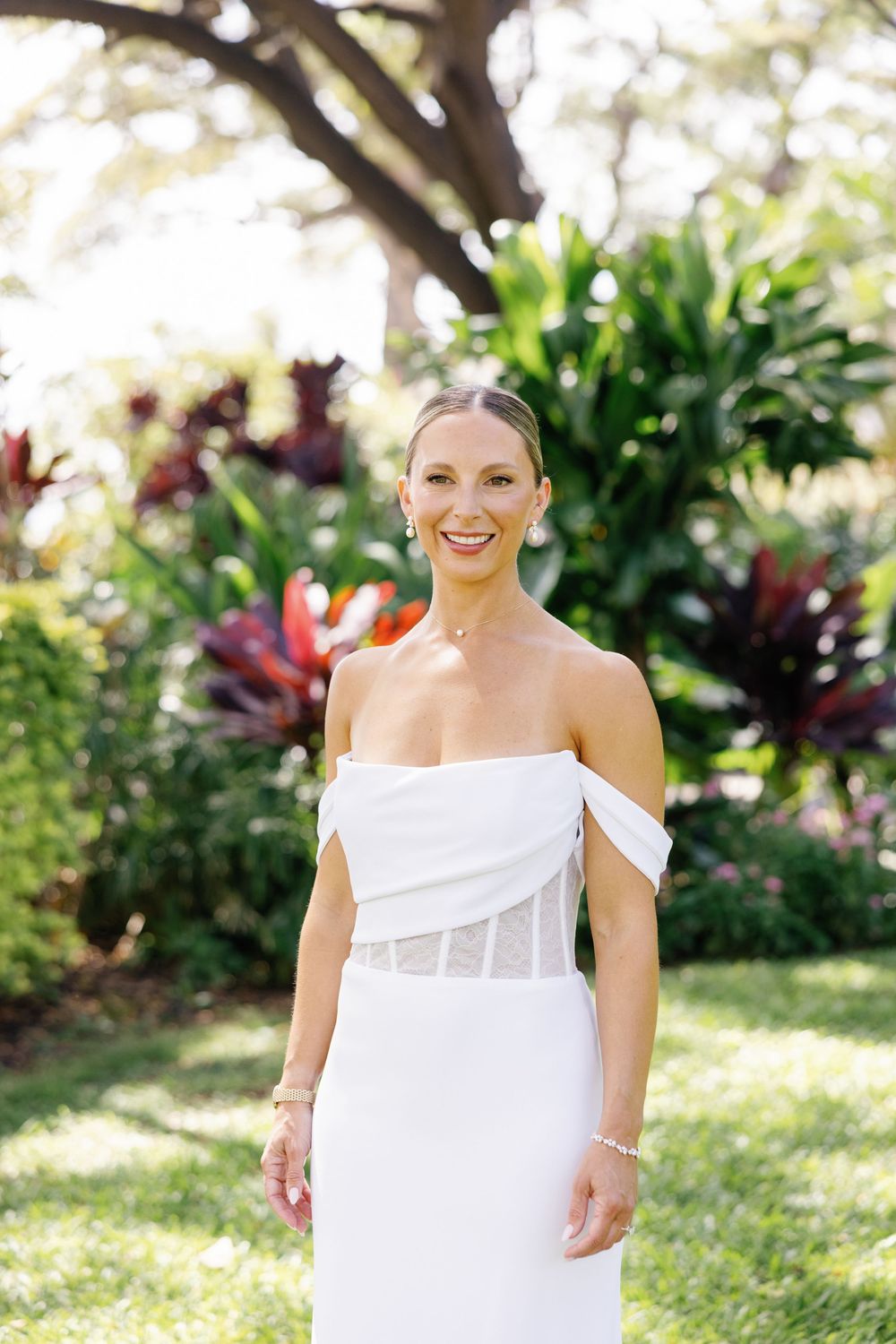 Smiling bride in white off-shoulder gown poses outdoors among tropical greenery and colorful foliage.
