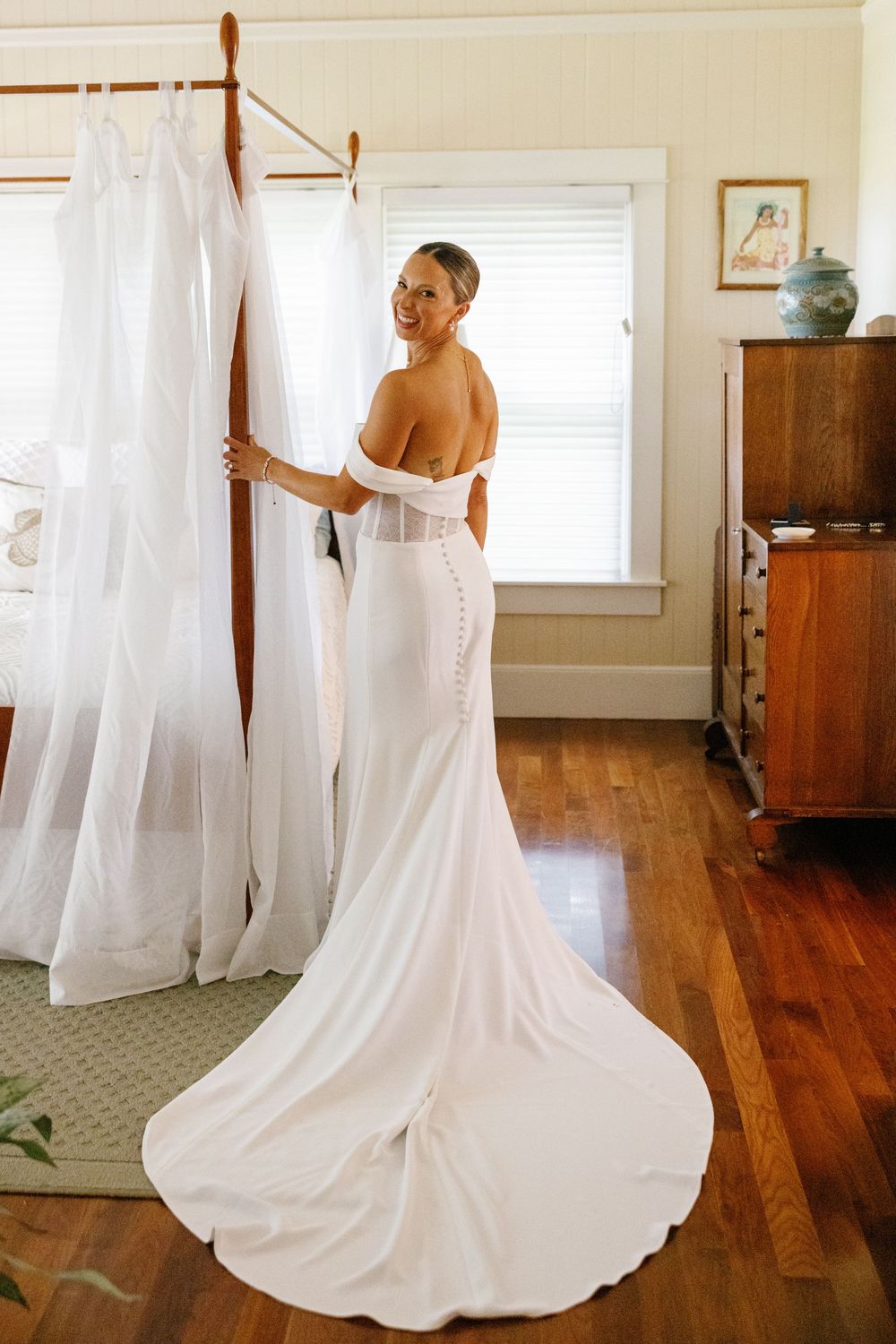 Bride in elegant off-shoulder white gown with button back train smiles in sunlit bedroom.