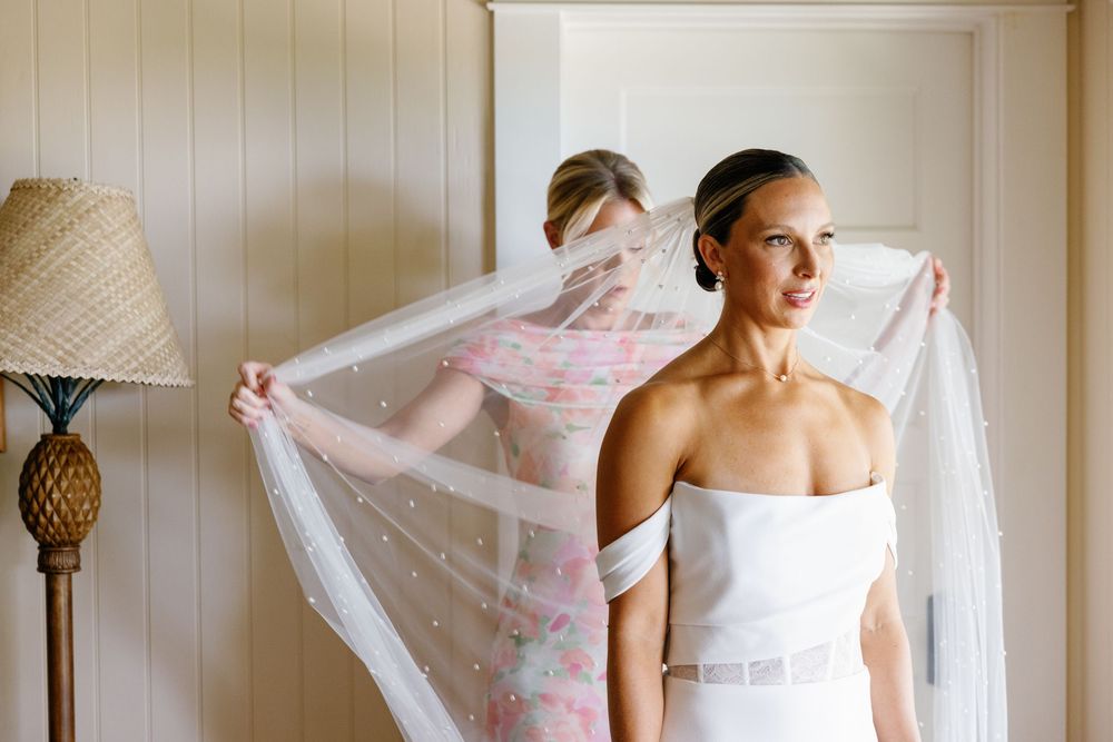 Bridesmaid in floral dress helps bride with pearl-embellished veil in bright white dressing room.