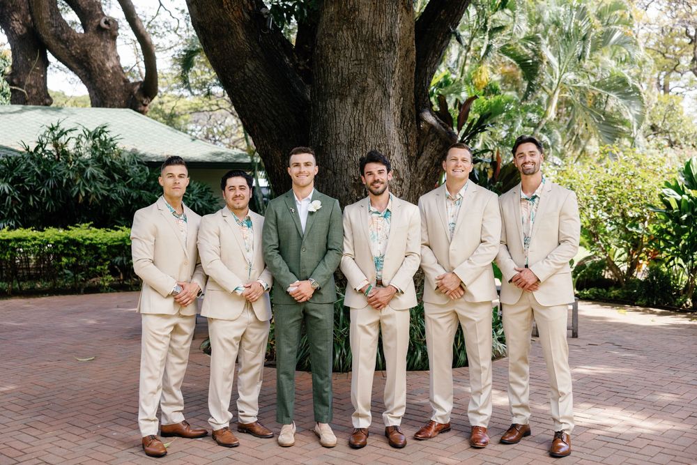 Groom in green suit poses with six groomsmen in tan suits outdoors near a large tree on a brick pathway.