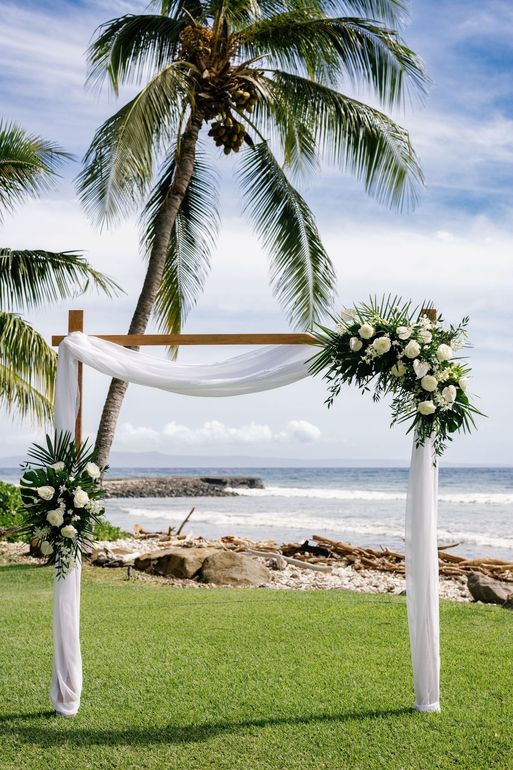Elegant beach wedding arch draped in white fabric with white rose floral arrangements beside a tropical palm tree.