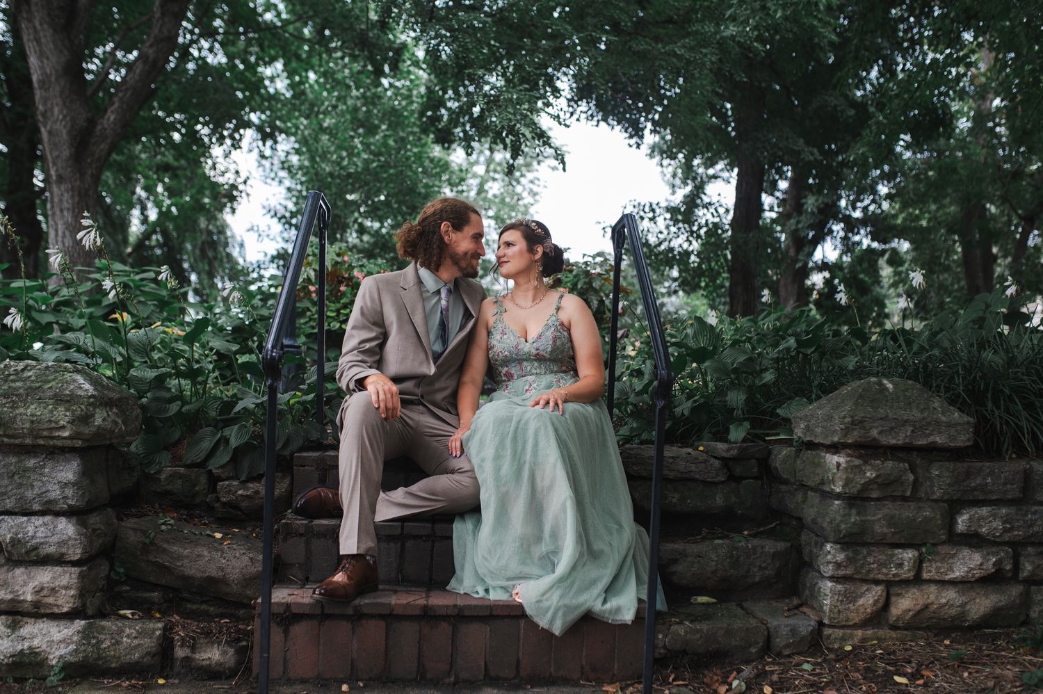 A couple in formal attire sits on stone steps surrounded by lush green trees, gazing lovingly at each other.