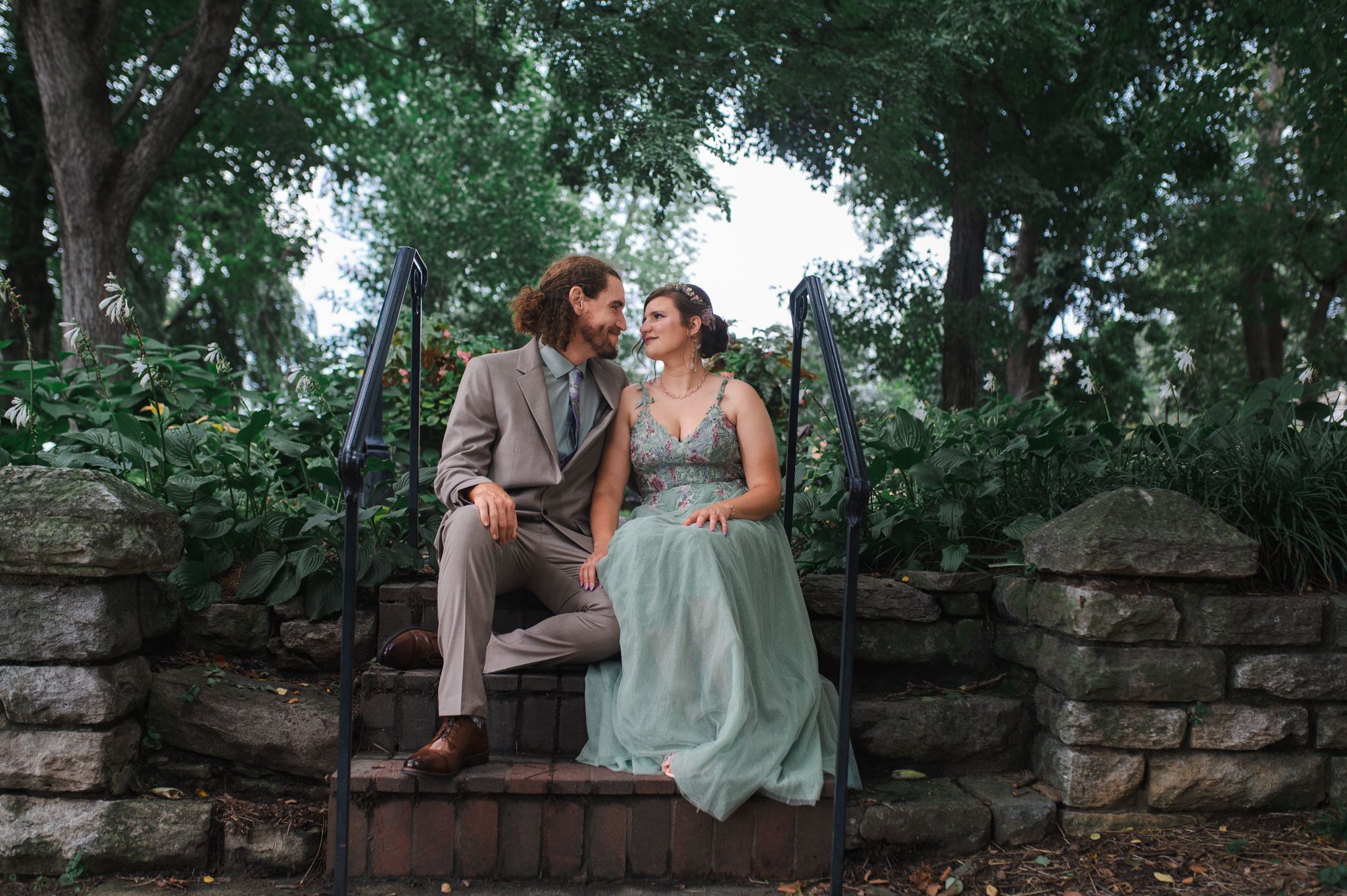 A couple in formal attire sits on stone steps surrounded by lush green trees, gazing lovingly at each other.