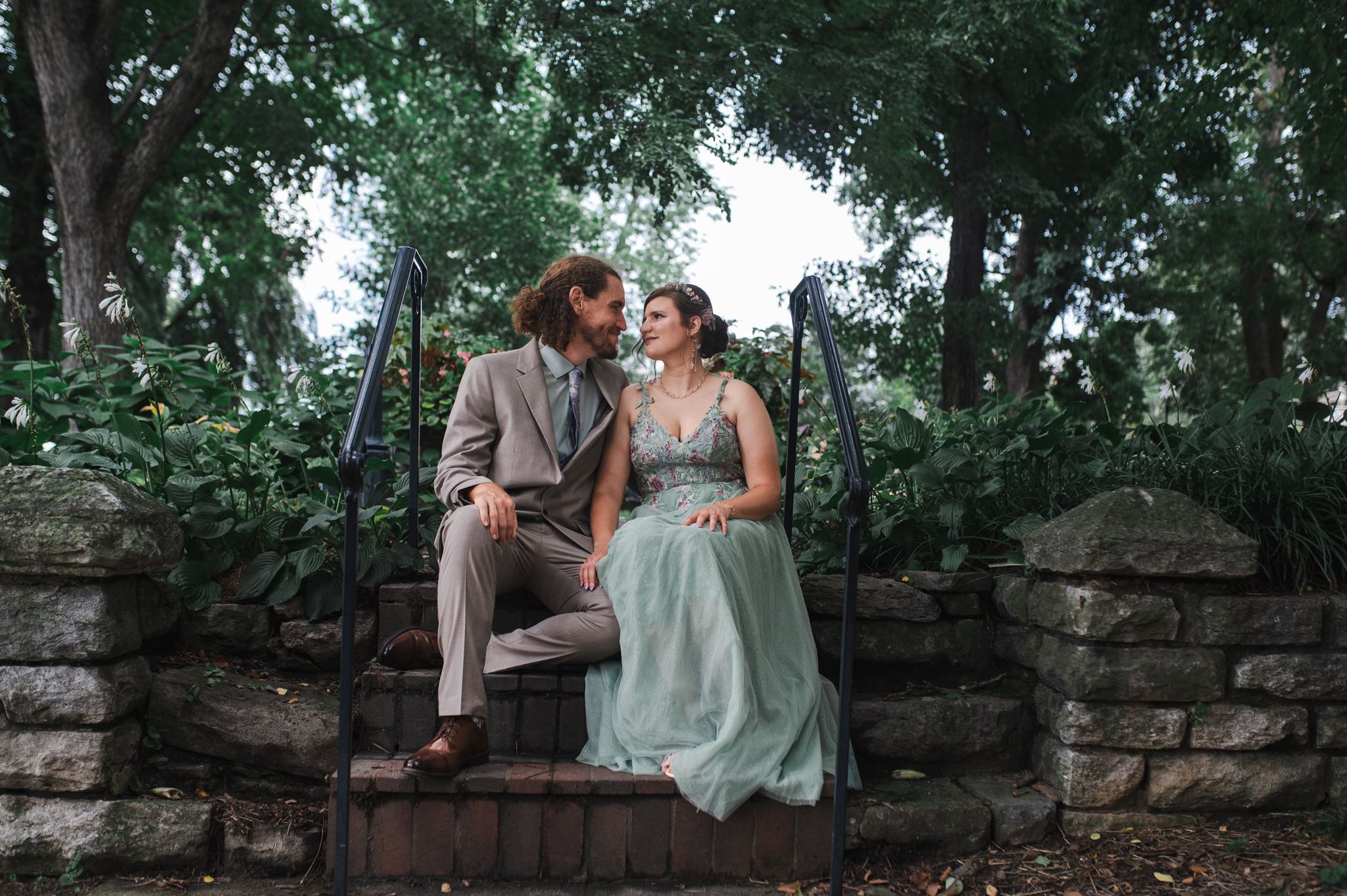 A couple in formal attire sits on stone steps surrounded by lush green trees, gazing lovingly at each other.