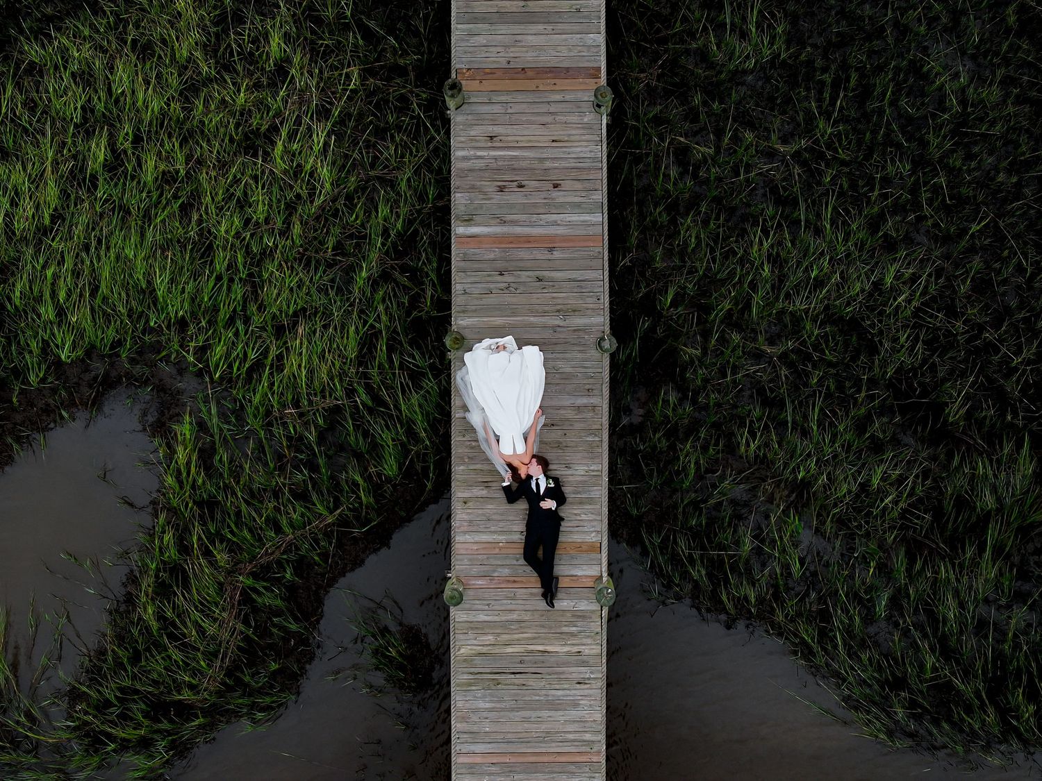 Aerial view of bride and groom lying on a wooden dock surrounded by lush green marshes and water.