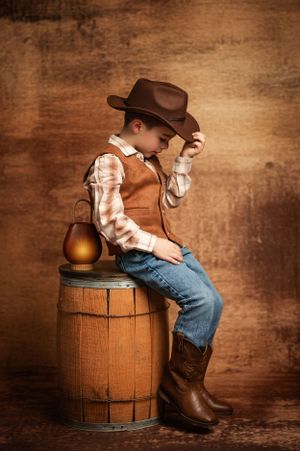 A western-themed portrait features a young cowboy in boots and hat sitting on a wooden barrel with an amber jug.