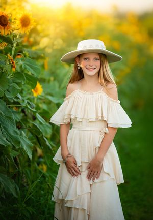 A white summer dress and sun hat stand out beautifully against a vibrant sunflower field backdrop in golden sunlight.