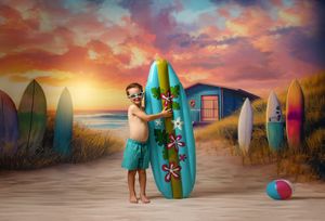 A young boy stands with a blue surfboard on a beach at sunset surrounded by colorful surfboards in the background.