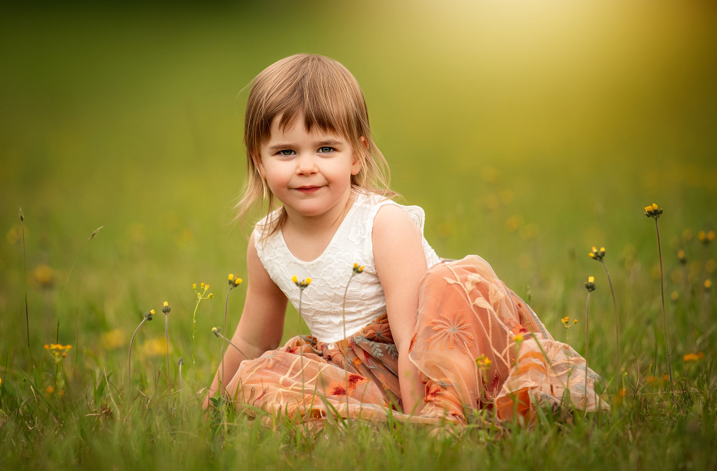 A smiling toddler girl in an orange floral dress sits in a sunny green meadow with yellow wildflowers.