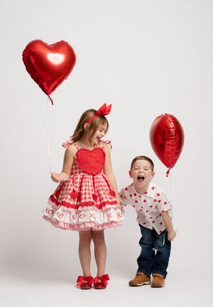 Two children in Valentine's themed outfits pose playfully with red heart balloons against a white studio backdrop.