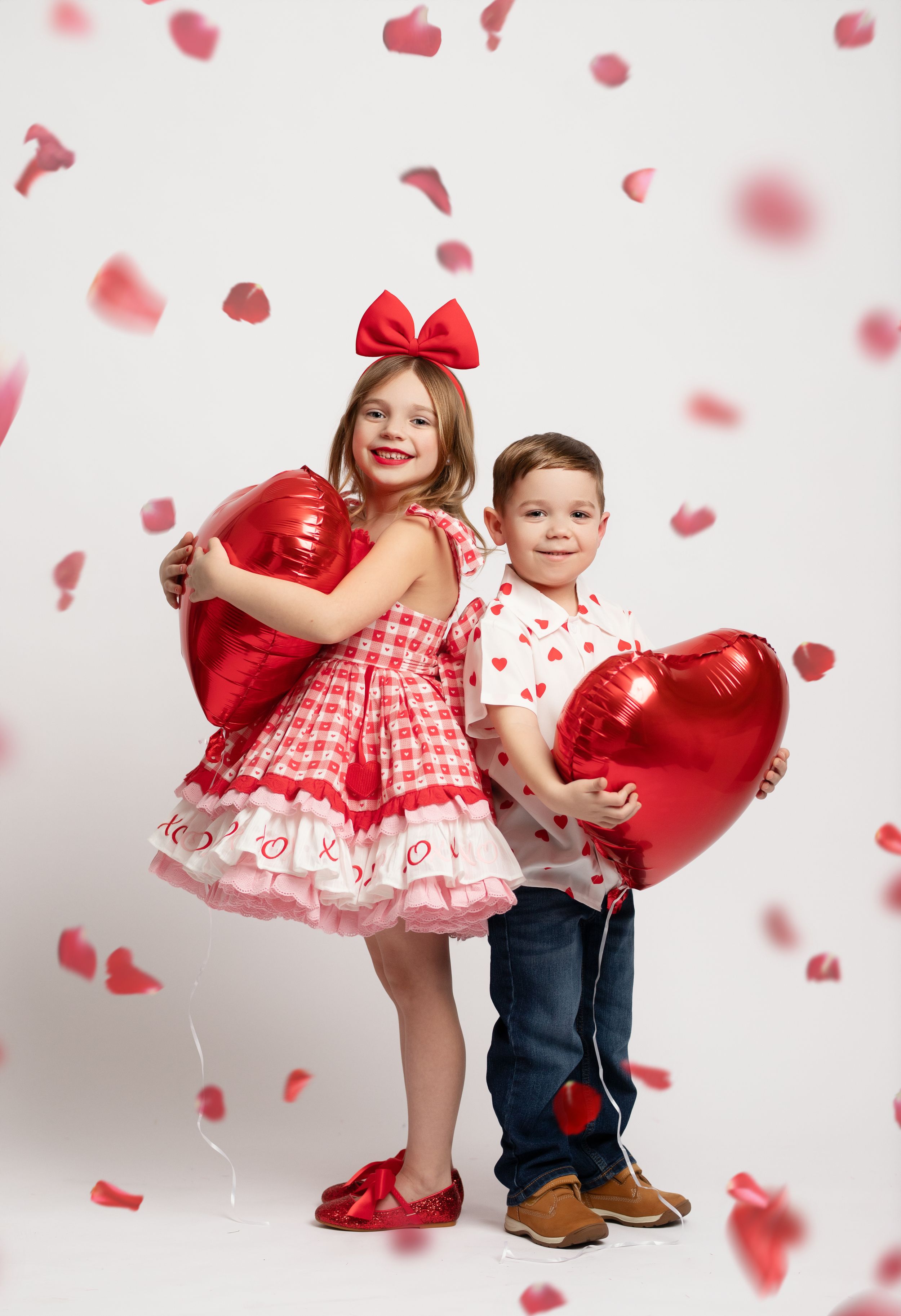 Children in red and white Valentine's Day outfits pose together as heart-shaped petals fall around them in a studio setting.