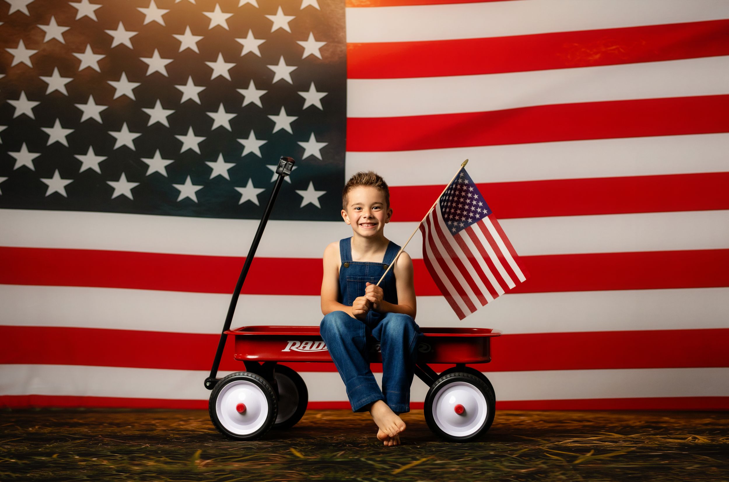 Young boy sitting in red Radio Flyer wagon holding American flag in front of large US flag backdrop.