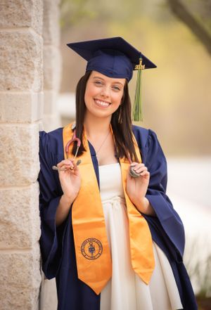 Graduate in navy cap and gown with gold sash celebrating on campus.
