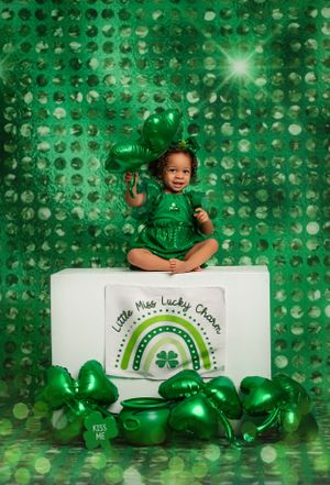 St Patrick's Day themed photo with sparkly green backdrop, rainbow sign, and green metallic heart balloons on white platform.
