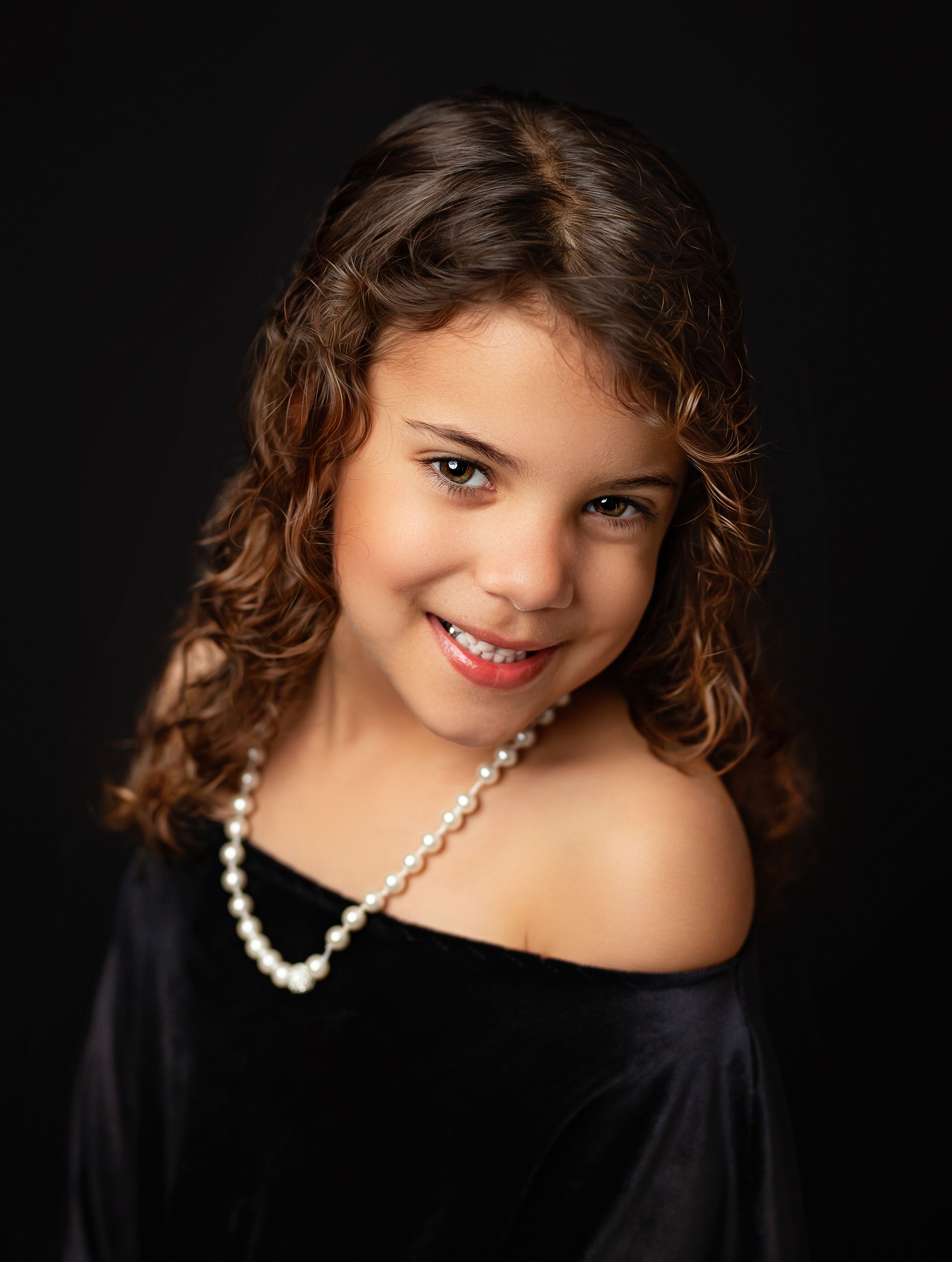A young girl with curly brown hair wearing a black off-shoulder top and pearl necklace smiles against a dark background.