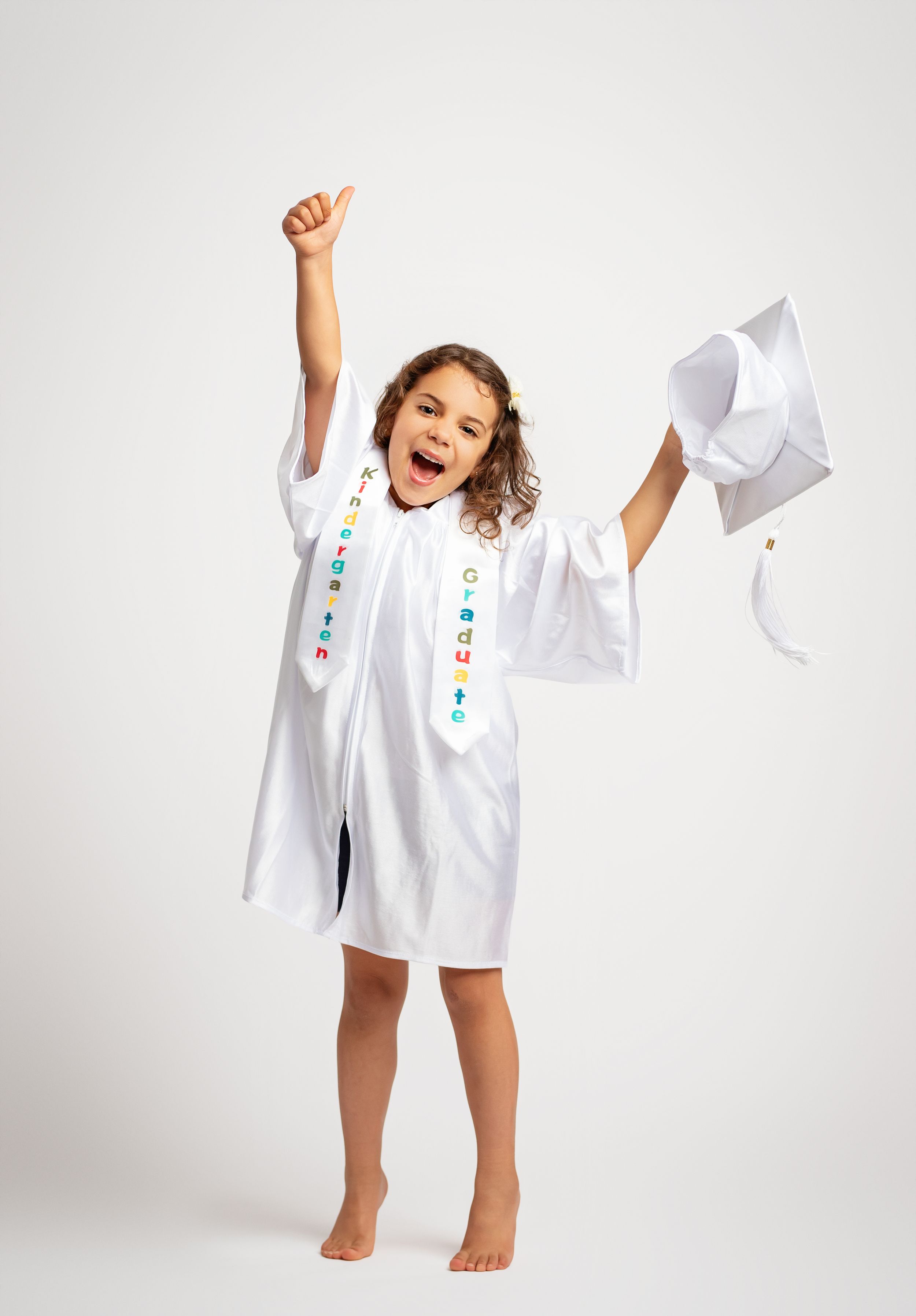 Excited young girl in white graduation gown and sash celebrating, holding cap with thumb up.