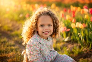 Young child with curly hair sits among colorful tulips in a garden during golden hour sunset lighting.