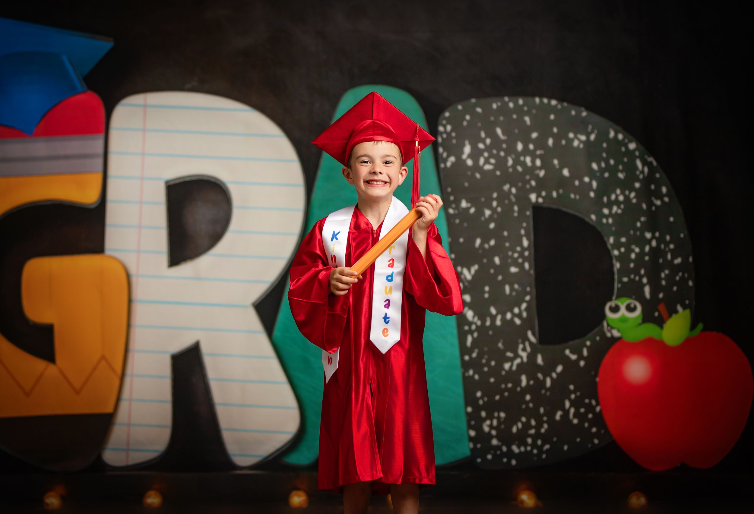 A small child dressed in red graduation cap and gown stands in front of large alphabet letters and apple classroom decoration.