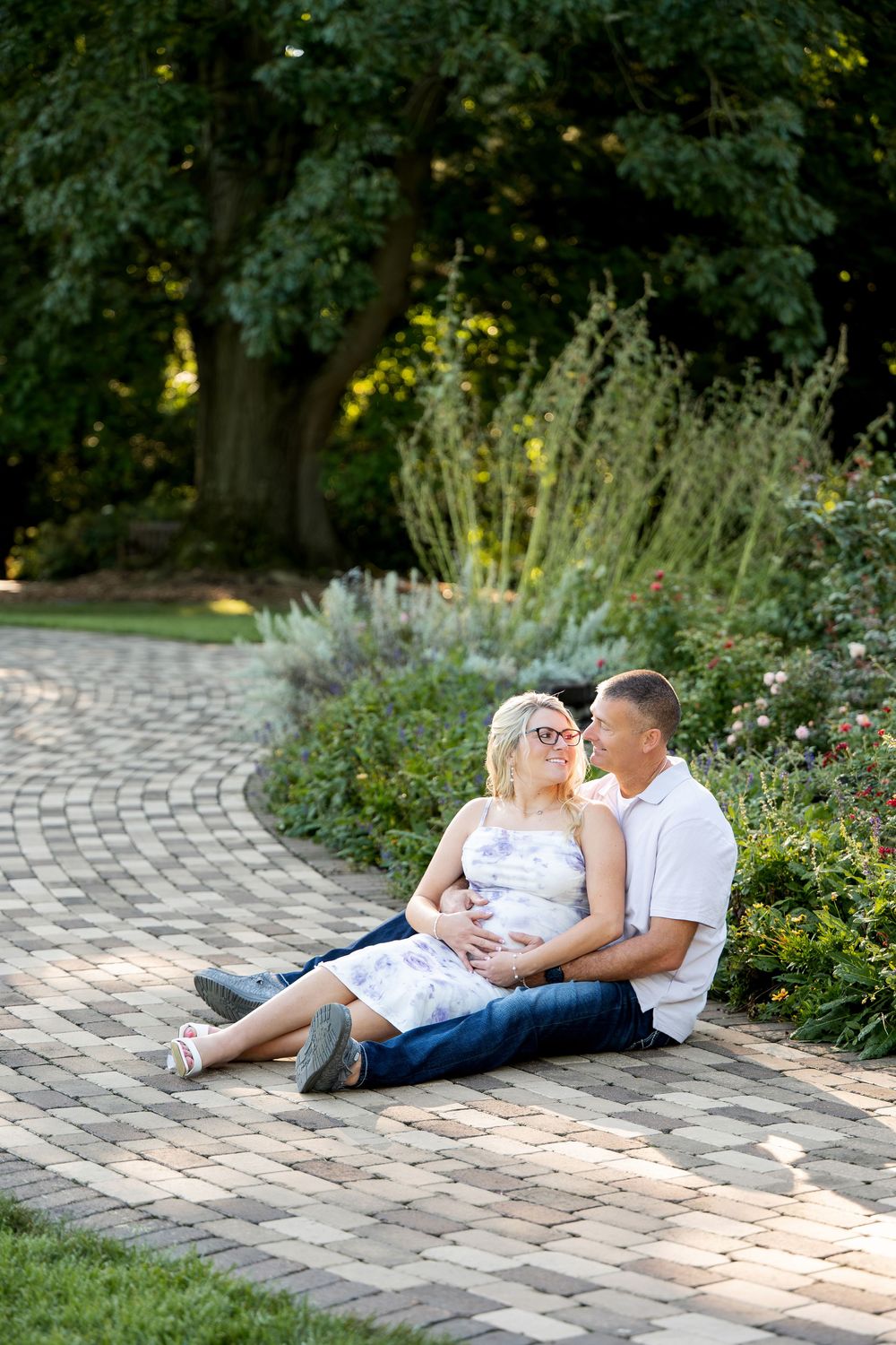 Couple sitting on brick path in garden, man embracing pregnant woman in floral dress, surrounded by lush greenery.