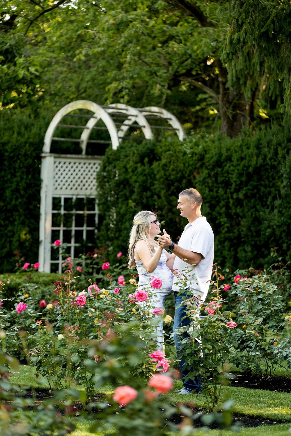 Couple sharing a romantic moment among colorful roses in a lush garden with a white arbor backdrop.