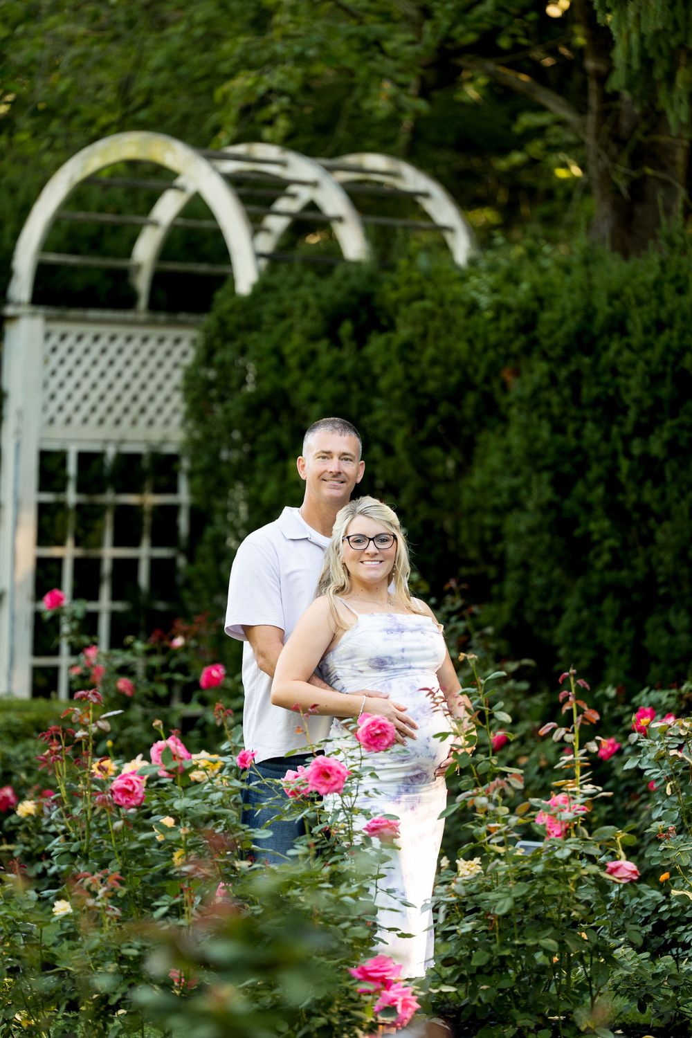 Couple posing in a rose garden with a white lattice arbor, woman in floral dress cradling baby bump.