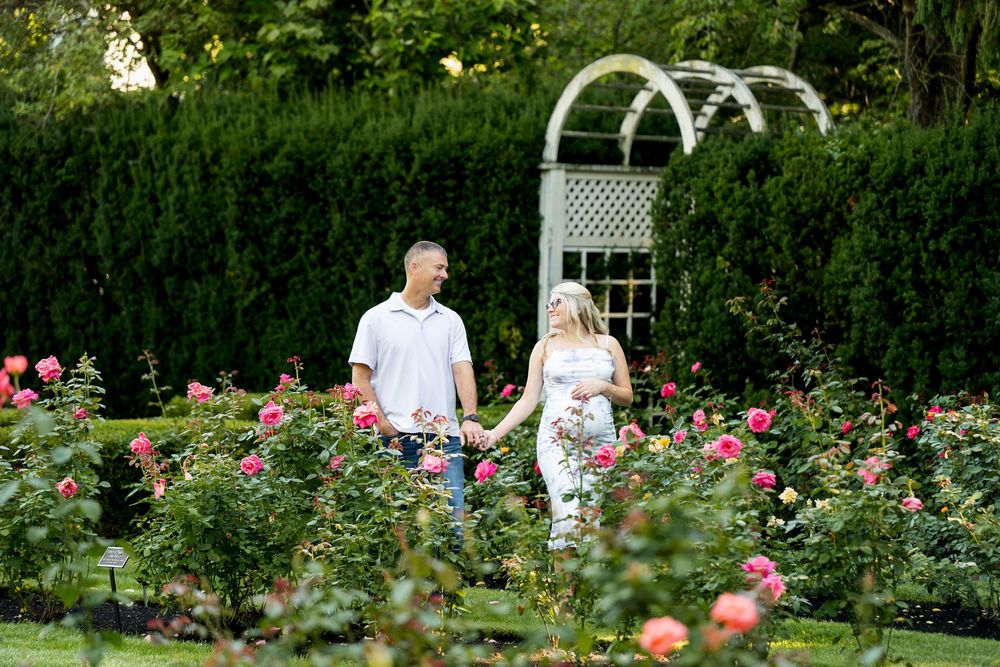 Couple holding hands walking through a lush rose garden with a white arbor backdrop.