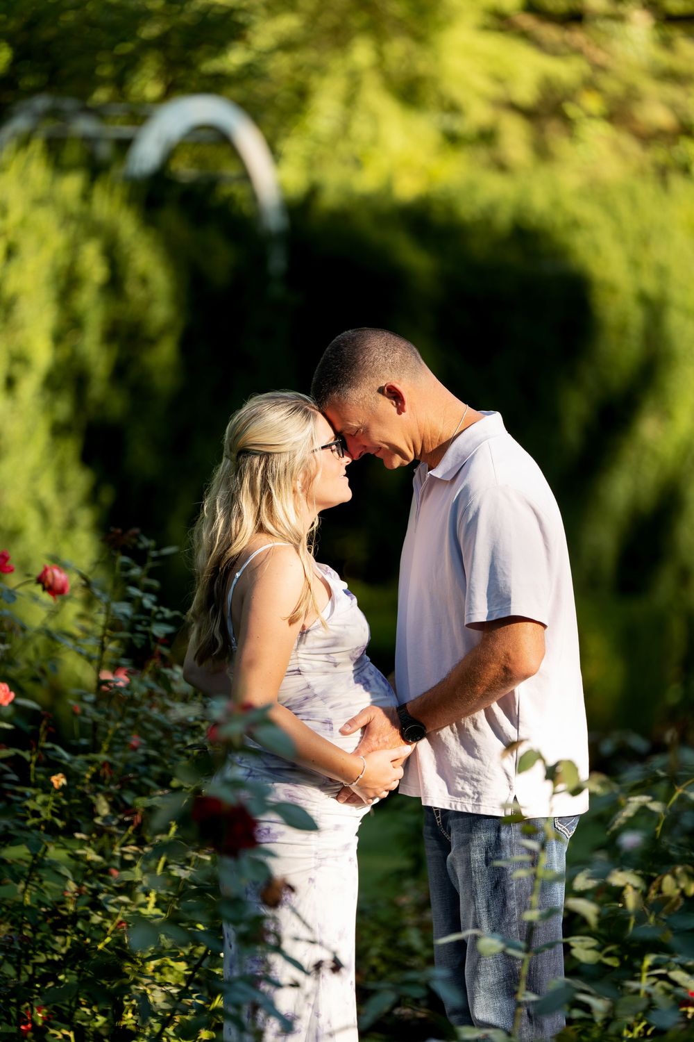 Expectant couple sharing an intimate moment in a rose garden, touching foreheads with hands on baby bump.
