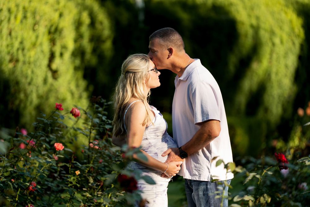Pregnant couple sharing a romantic moment in a rose garden, surrounded by lush green hedges.