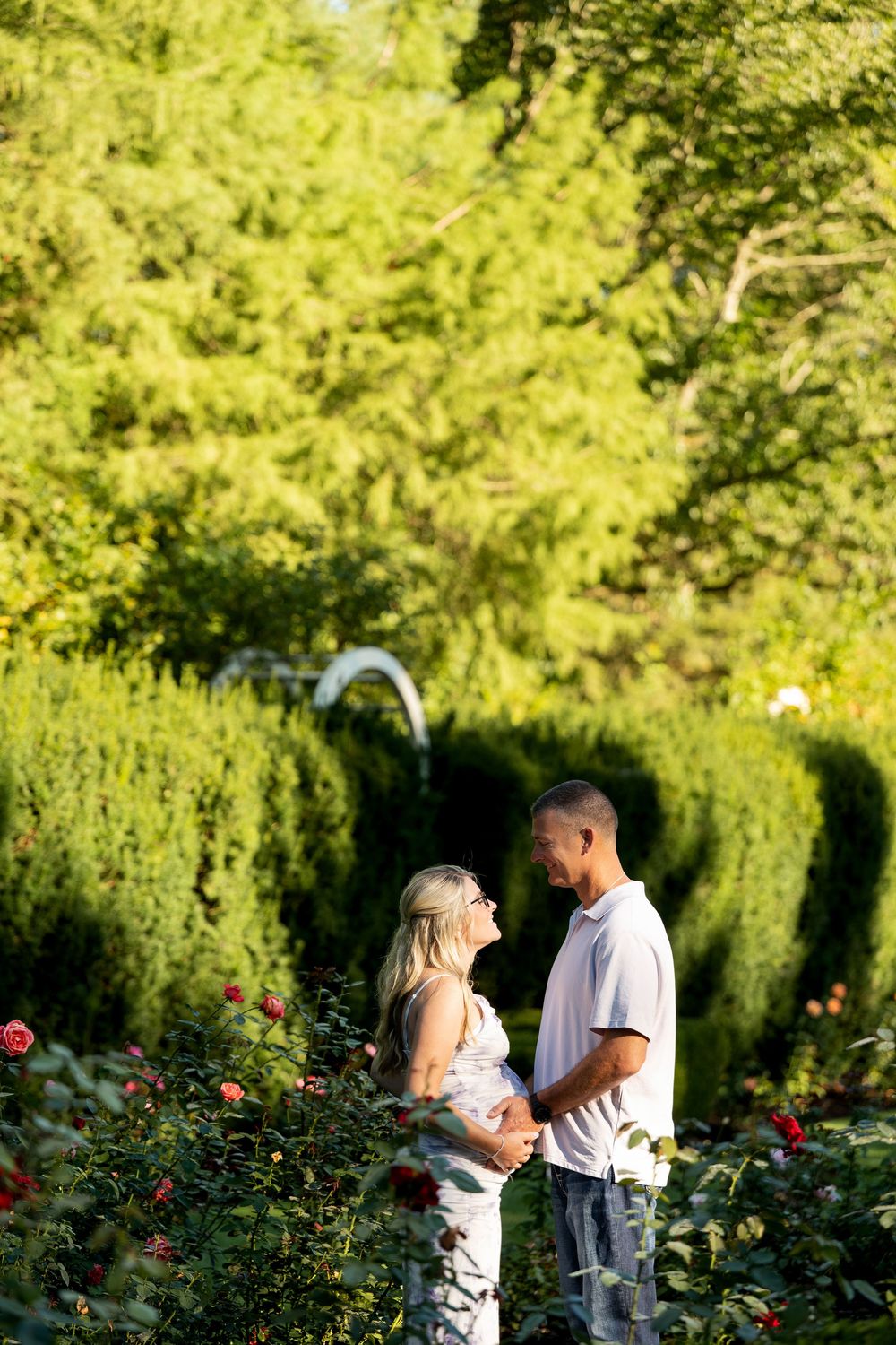 Couple standing together among red roses in a lush green garden, smiling at each other on a sunny day.