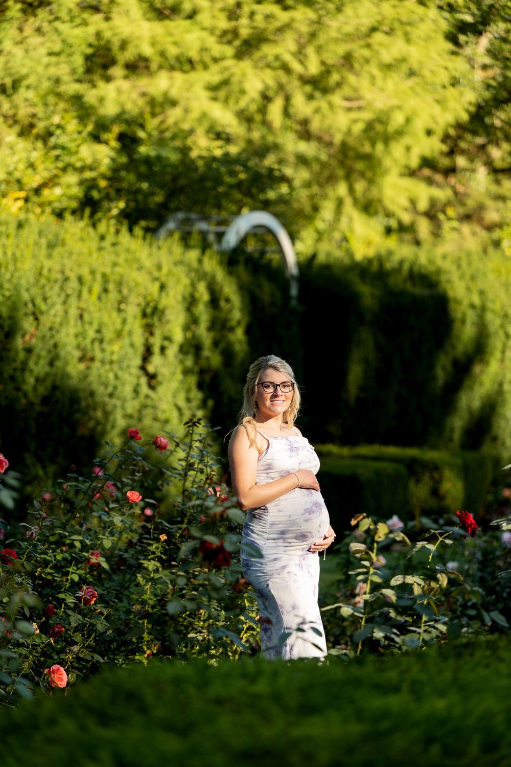 Smiling pregnant woman in a floral dress cradling her baby bump while standing among red roses in a garden.
