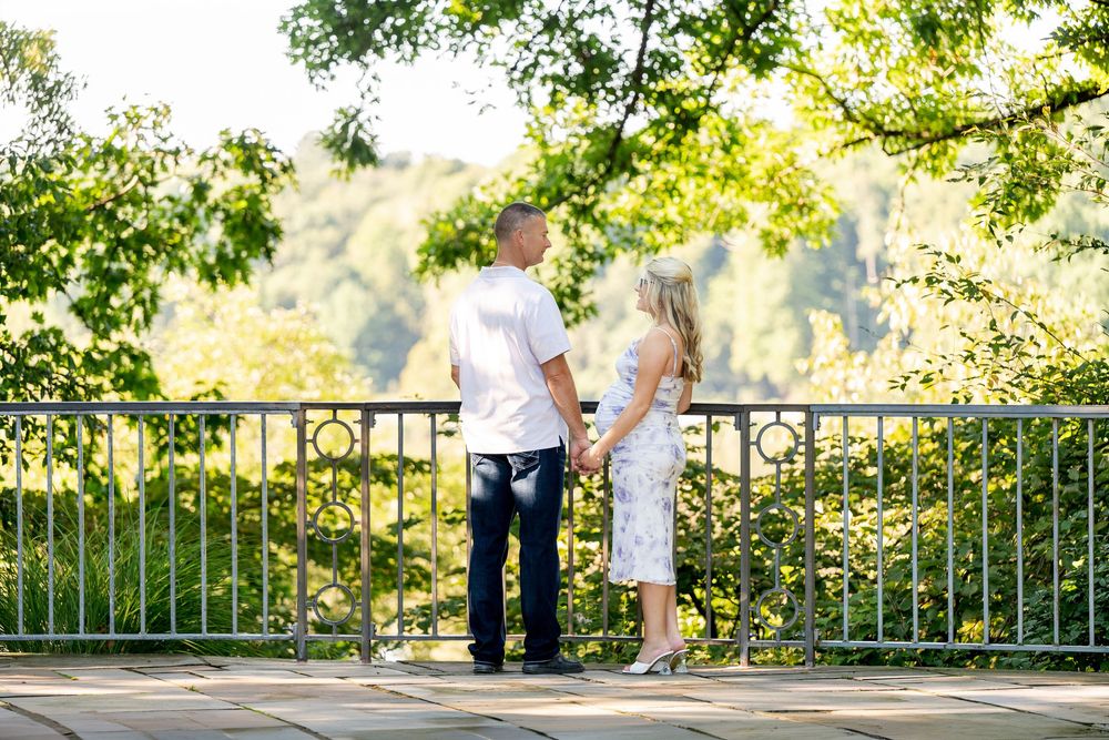 Couple holding hands on a scenic bridge overlook surrounded by lush green trees on a sunny day.