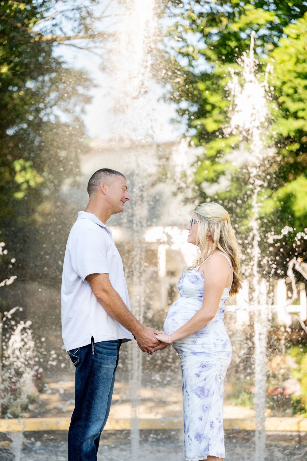 Expecting couple holding hands and gazing at each other in front of a beautiful garden fountain on a sunny day.