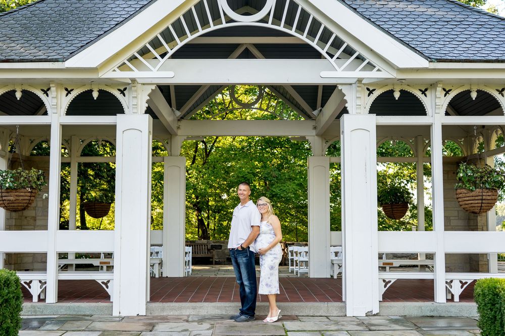 Pregnant couple embracing and smiling together under a white gazebo with hanging flower baskets in a garden.