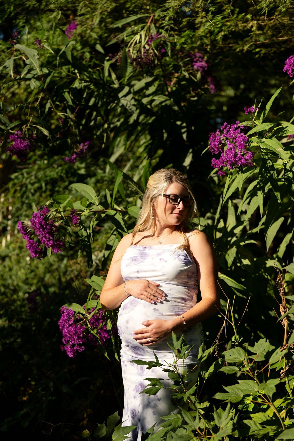 Pregnant woman in floral dress cradling her bump among vibrant purple lilac bushes in sunlight.