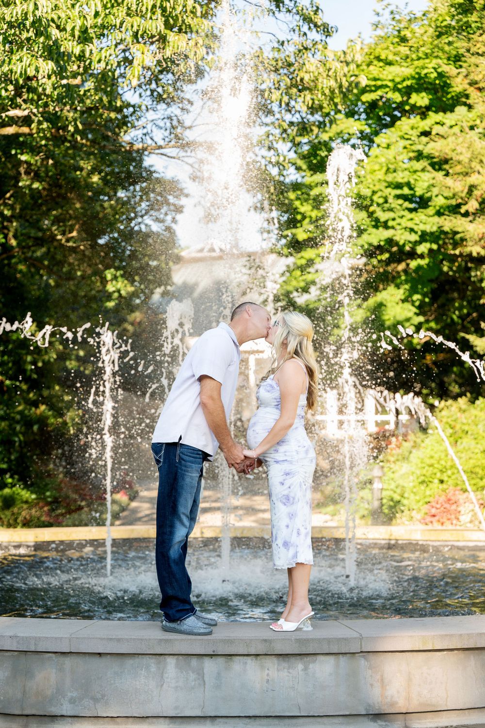 Couple kissing in front of a garden fountain, woman in floral dress holding hands with man in white shirt and jeans.