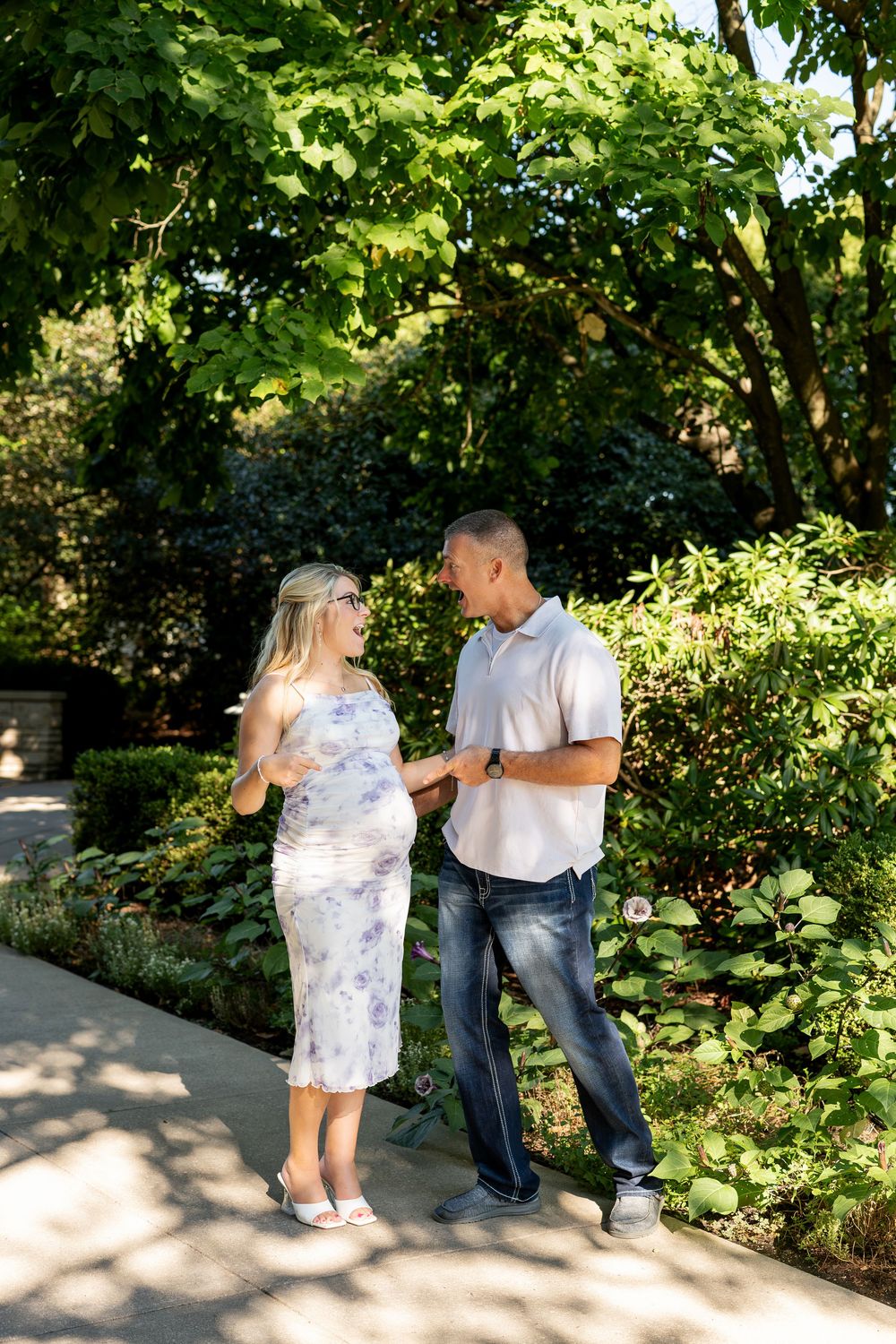 Pregnant couple laughing together on a garden path surrounded by lush green trees and shrubs on a sunny day.