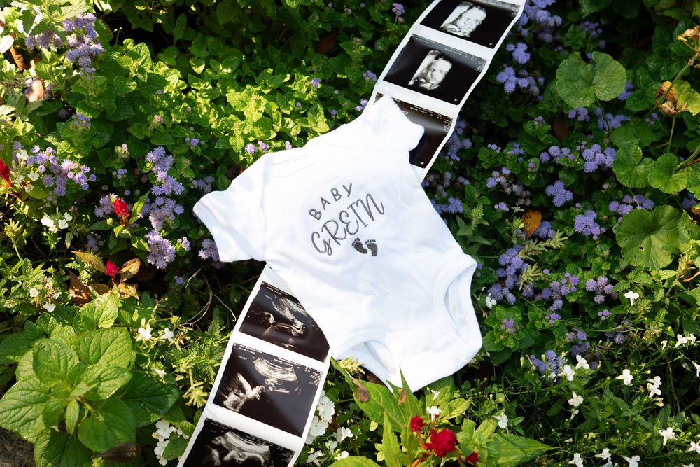 Baby onesie with ultrasound photos on green floral background for pregnancy announcement.