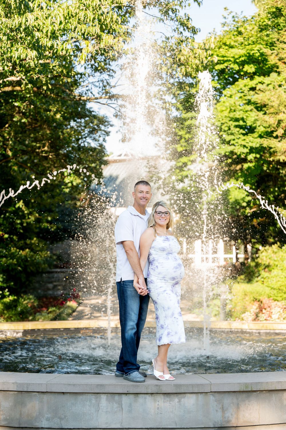 Pregnant couple holding hands smiling in front of a garden fountain surrounded by lush green trees.