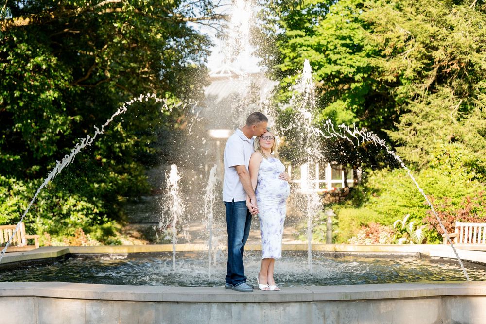 Couple embracing in front of a fountain in a lush garden, surrounded by greenery and sunlight.