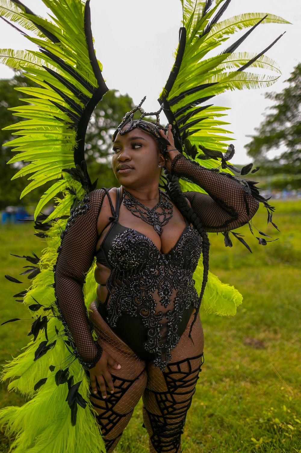Woman in vibrant green and black carnival costume with large feathered wings poses outdoors at a festival.