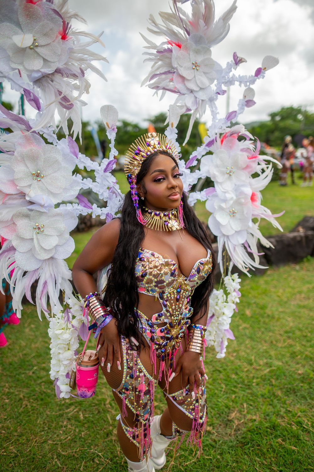 Woman in colorful carnival costume with white floral wings and gold crown poses outdoors at a festival.