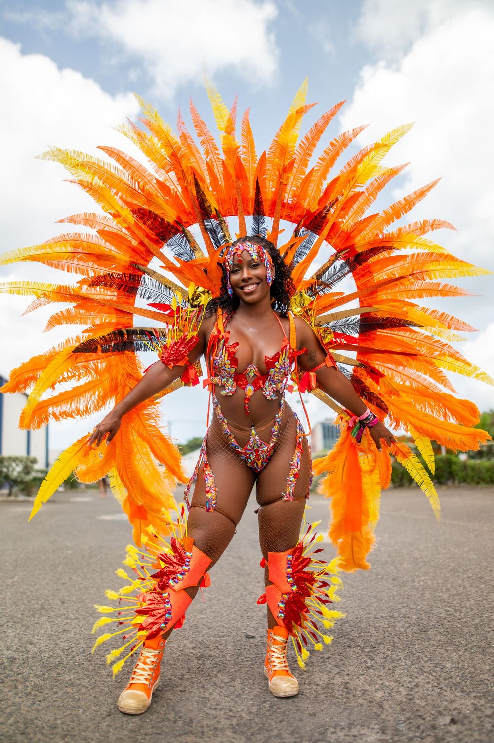 Woman in vibrant orange feathered carnival costume smiling and posing outdoors at a festive parade.