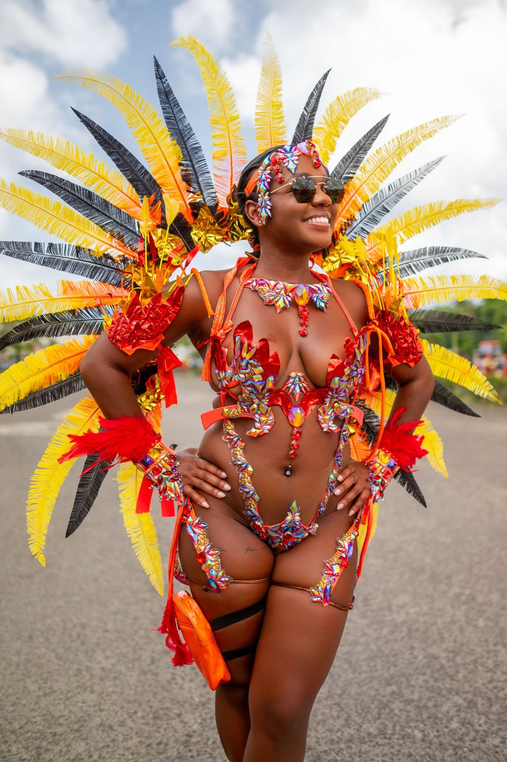Woman in vibrant carnival costume with yellow and black feathers, orange bikini, and jeweled accessories poses outdoors.
