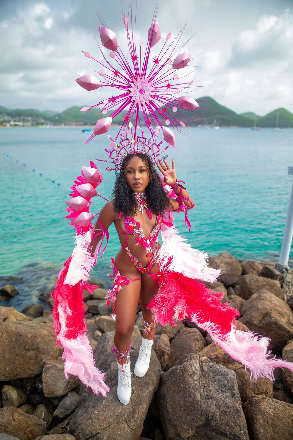 Woman in vibrant pink carnival costume with feathers and headdress posing on rocks by turquoise Caribbean sea.