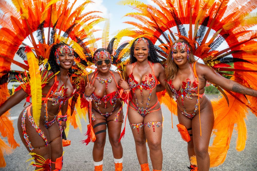 Four women in vibrant orange feathered carnival costumes pose together at a colorful festival parade.