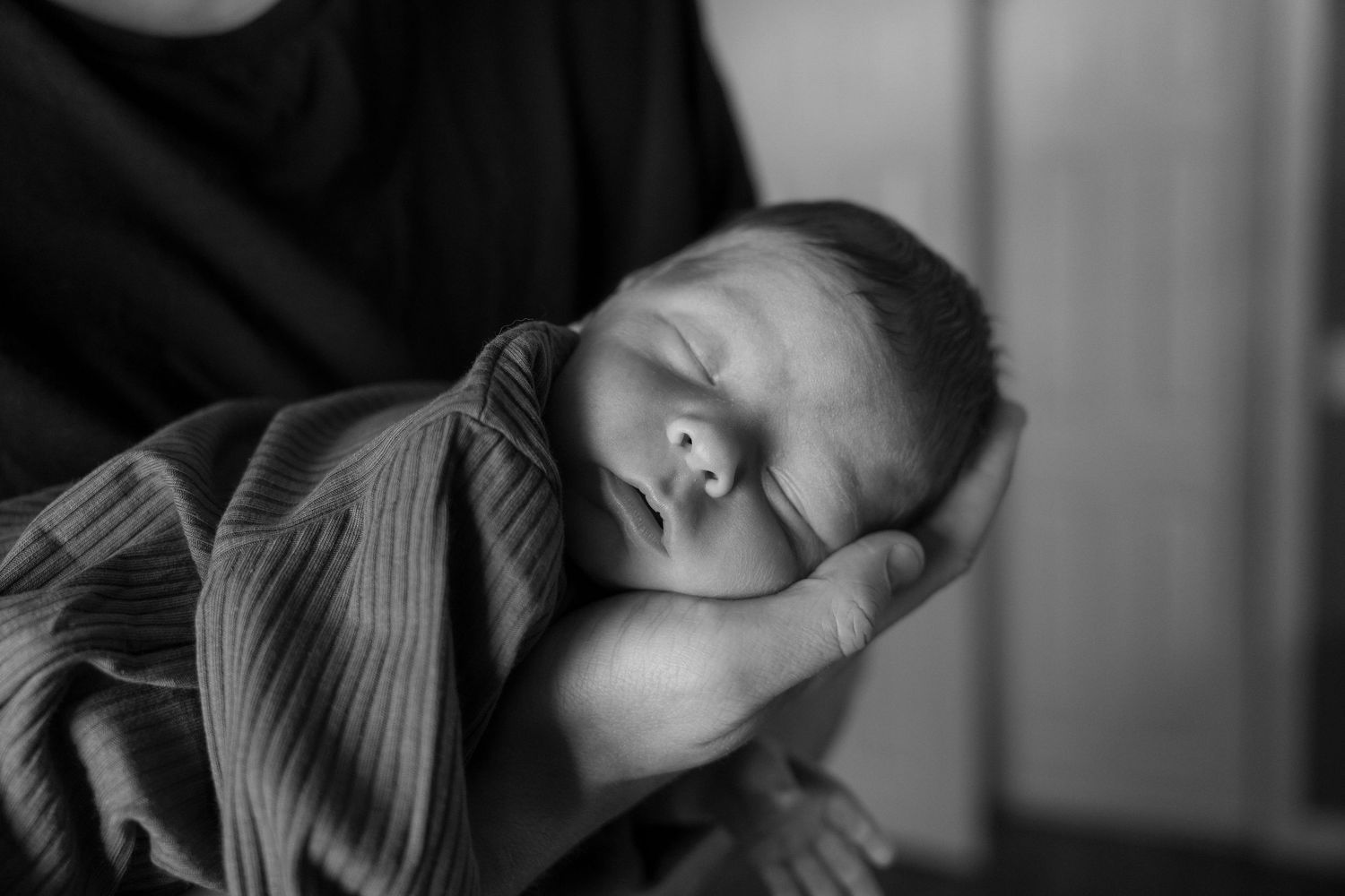 Black and white photo of a sleeping newborn baby wrapped in a swaddle, cradled gently in an adult's hands.