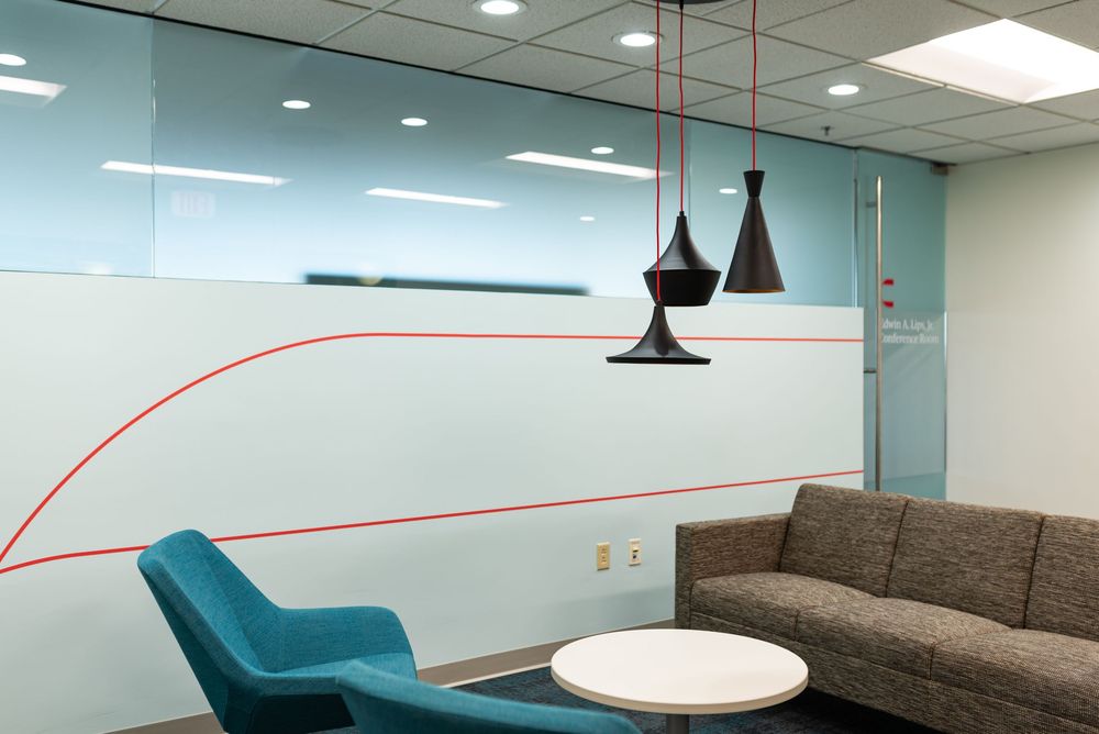 Modern office lounge with teal chairs, gray sofa, white table, black pendant lights, and red-lined frosted glass wall.