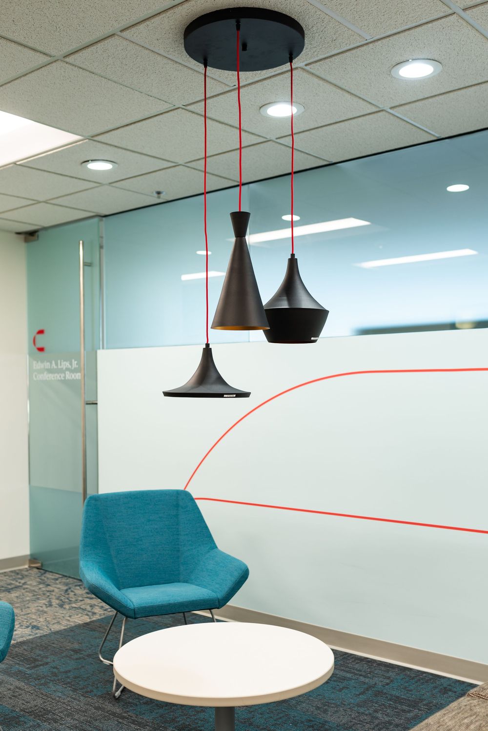 Modern office lounge with teal chair, round table, and three black pendant lights with red cords near frosted glass wall.